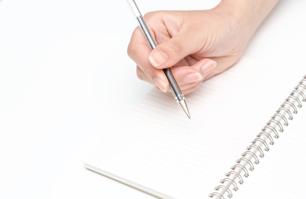 ノートにペンで書き込む手元。見つけた表記を書き写して見比べるイメージ(実際に選んだ画像に合わせて直す)